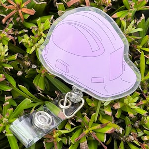 Puede incluir: Un portatarjetas con una ilustración de un casco de seguridad de color violeta claro encapsulado en acrílico transparente. El diseño del casco incluye detalles de líneas negras. El portatarjetas tiene una correa de plástico transparente.