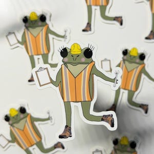 May include: Sticker sheet featuring cartoon frogs in construction attire. Each frog wears a yellow hard hat and an orange safety vest with reflective stripes. Some frogs hold clipboards, others hold wrenches.