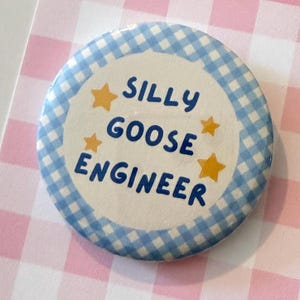 May include: A round pinback button featuring a blue and white gingham border. The center is white with the text SILLY GOOSE ENGINEER printed in dark blue capital letters, accented by four small yellow stars arranged around the text.