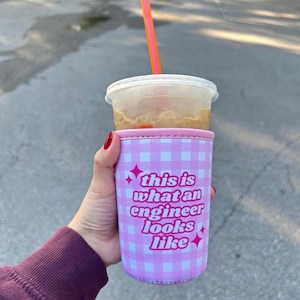 Puede incluir: Una funda de neopreno para vaso con estampado de cuadros vichy en rosa claro y blanco, con el texto en rosa oscuro "this is what an engineer looks like" y estrellas, alrededor de un vaso de plástico transparente con café helado y pajita naranja.