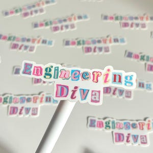 May include: A collection of stickers with the text "Engineering Diva" in a colorful, cut-out style. The stickers feature pink, blue, and yellow hues. One sticker is held by a white pen. The stickers are scattered across a white surface.