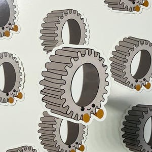 May include: A collection of stickers featuring a cartoon gear design. The gears are gray with a smiling face and small brown feet. The stickers are on a white background.