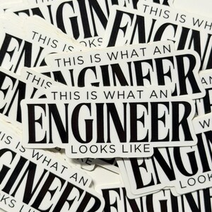 May include: A collection of white rectangular stickers with black text. The stickers read "THIS IS WHAT AN ENGINEER LOOKS LIKE" in a bold, sans-serif font. The stickers are scattered across a white surface.
