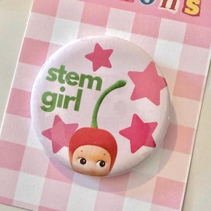 May include: A circular pinback button featuring a light pink background with four dark pink stars. The text 'stem girl' is printed in green letters next to a graphic of a red cherry with a face, featuring large eyes and a small mouth.