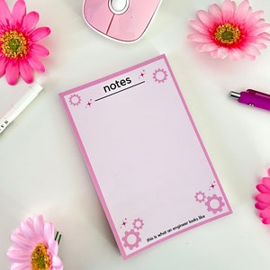 May include: Pink notepad with the word "notes" written on it. The notepad has a pink border and a white interior. The notepad is decorated with small gears and stars. The text "this is what an engineer looks like" is written at the bottom of the notepad.