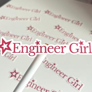 May include: A white sticker with the text "Engineer Girl" in a bold, red font, accented with red star shapes. The sticker is held by a white stick, with other stickers in the background.