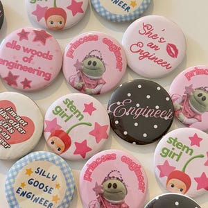 May include: A collection of circular pin-back buttons featuring various designs. Some buttons display text such as 'elle woods of engineering', 'She's an Engineer', 'stem girl', 'silly goose engineer', and 'princess engineer' alongside illustrations of cherries, stars, and a frog.