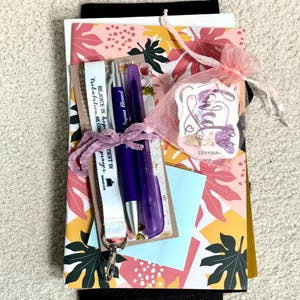 May include: A stationery set featuring a floral patterned card, a purple pen, a purple highlighter, a white lanyard with the text REJOICE IN hope TIENT IN prayer, and a pink organza bag containing a sticker with the word Loved and floral art.