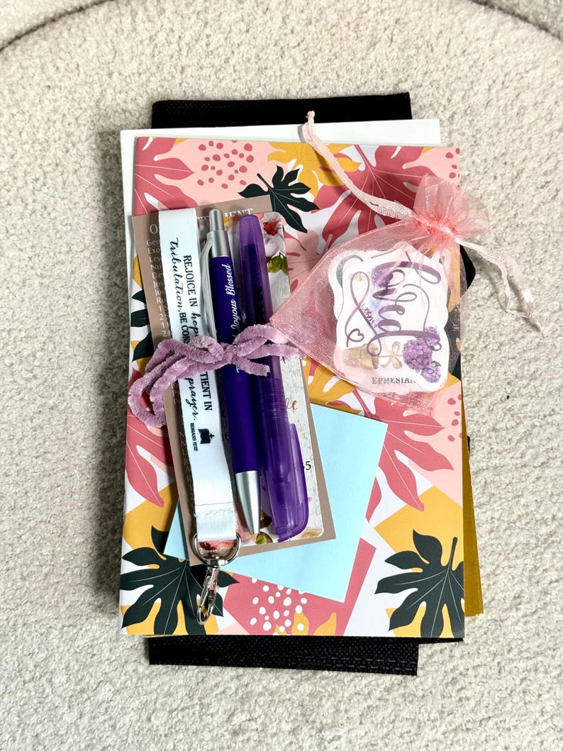 May include: A stationery set featuring a floral patterned card, a purple pen, a purple highlighter, a white lanyard with the text REJOICE IN hope TIENT IN prayer, and a pink organza bag containing a sticker with the word Loved and floral art.