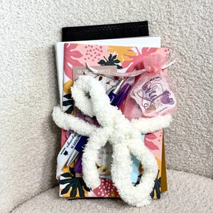 May include: A stack of notebooks and papers featuring a pink, yellow, and green leaf pattern, secured with a white fuzzy yarn bow. Included are two pens, a small pink organza drawstring bag with a tag reading "Loved," and a black folder.