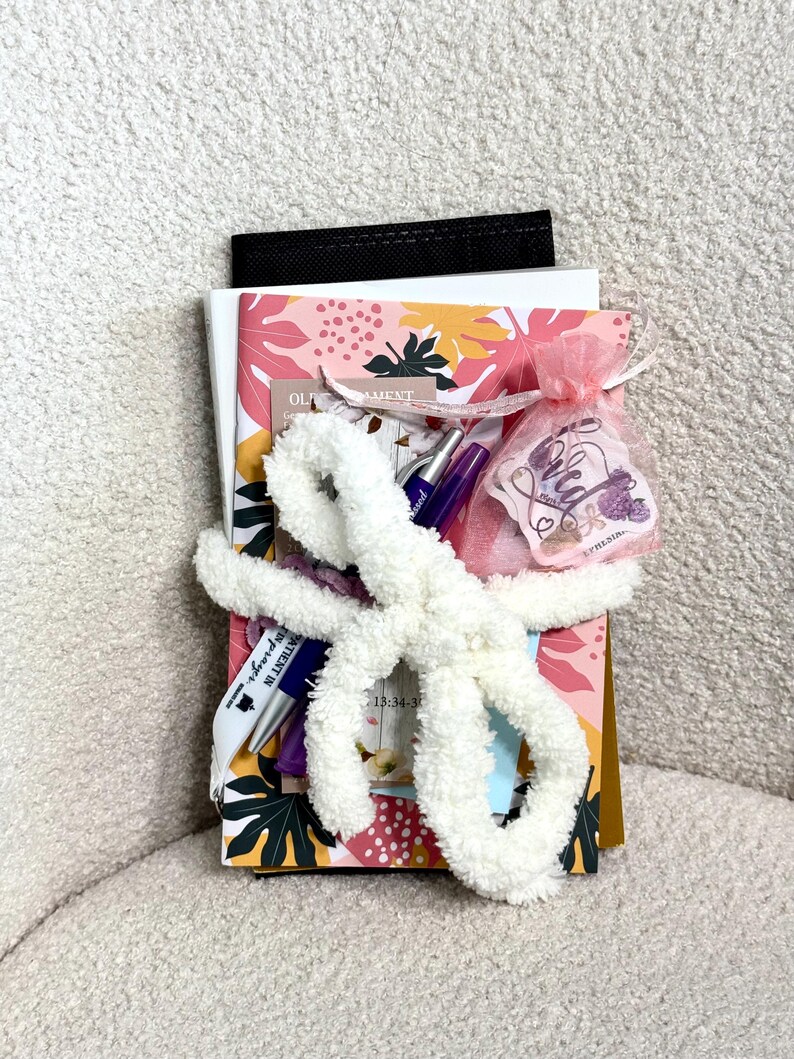 May include: A stack of notebooks and papers featuring a pink, yellow, and green leaf pattern, secured with a white fuzzy yarn bow. Included are two pens, a small pink organza drawstring bag with a tag reading "Loved," and a black folder.