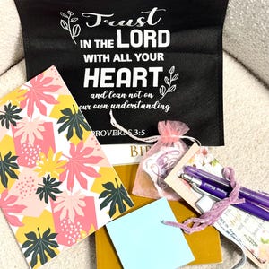 May include: A black tote bag with white text reading Trust in the Lord with all your heart and lean not on your own understanding Proverbs 3:5, a notebook with a pink and yellow leaf pattern, a light blue notepad, a gold book, and two purple pens.