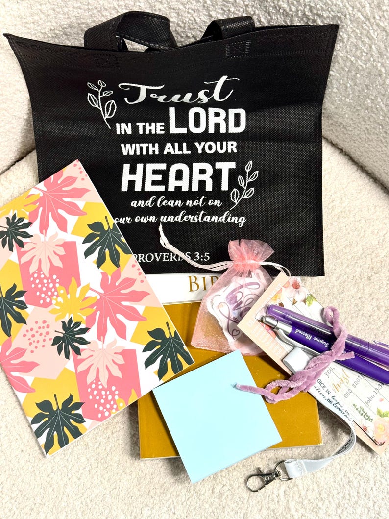 May include: A black tote bag with white text reading Trust in the Lord with all your heart and lean not on your own understanding Proverbs 3:5, a notebook with a pink and yellow leaf pattern, a light blue notepad, a gold book, and two purple pens.