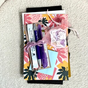 May include: A stack of items including a notebook with a pink, yellow, and dark green leaf pattern, a purple pen, a clear purple highlighter, a white lanyard with the text "REJOICE IN hope", and a pink organza bag containing a sticker labelled "loved".