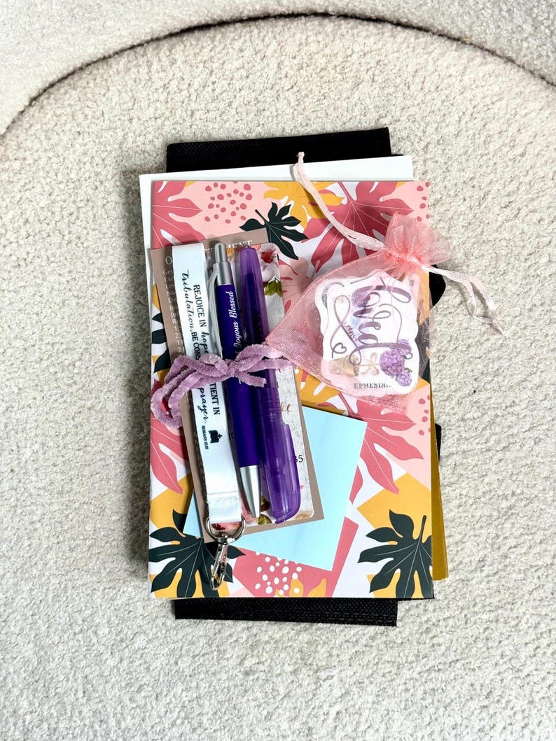 May include: A stack of items including a notebook with a pink, yellow, and dark green leaf pattern, a purple pen, a clear purple highlighter, a white lanyard with the text "REJOICE IN hope", and a pink organza bag containing a sticker labelled "loved".
