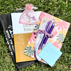May include: A Holy Bible with a gold and white cover, a floral patterned notebook, a pink organza bag containing a sticker, two purple pens, a white lanyard with text, and a light blue sticky note pad, all arranged together on a green grass surface.