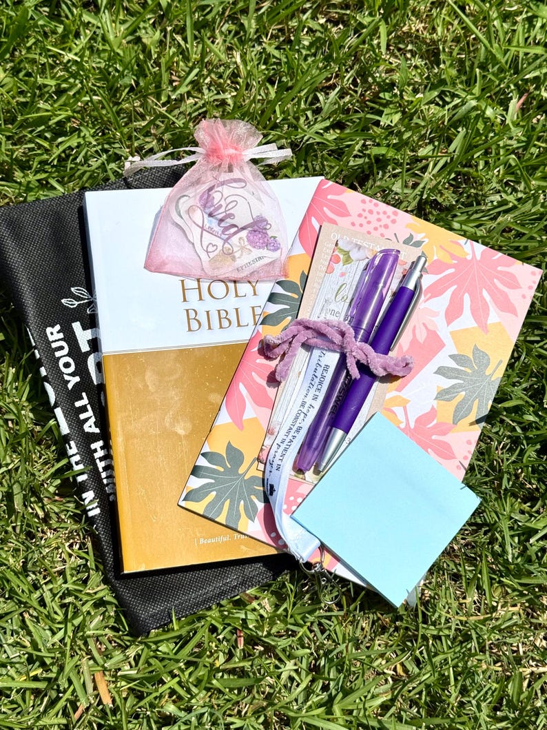 May include: A Holy Bible with a gold and white cover, a floral patterned notebook, a pink organza bag containing a sticker, two purple pens, a white lanyard with text, and a light blue sticky note pad, all arranged together on a green grass surface.