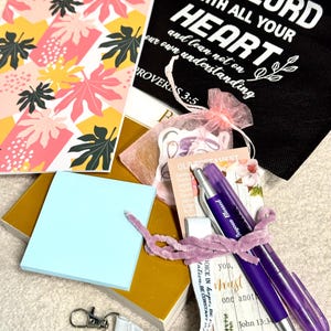 May include: A collection of stationery items including a notebook with pink and yellow leaf patterns, a black bag with white text reading Trust in the Lord, a mustard yellow book, a light blue sticky note pad, a purple pen, and a white lanyard.
