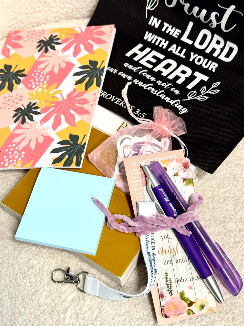 May include: A collection of stationery items including a notebook with pink and yellow leaf patterns, a black bag with white text reading Trust in the Lord, a mustard yellow book, a light blue sticky note pad, a purple pen, and a white lanyard.