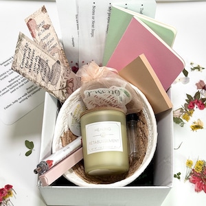 May include: A gift box filled with a woven basket containing a candle labeled "Healing Retablissement", small notebooks, bookmarks with text, a pen, and a small vial. The box is white, and the contents are arranged with dried flowers.