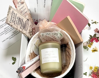 May include: A gift box filled with a woven basket containing a candle labeled "Healing Retablissement", small notebooks, bookmarks with text, a pen, and a small vial. The box is white, and the contents are arranged with dried flowers.