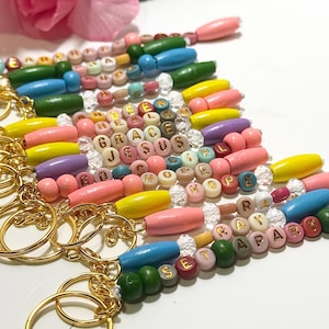 May include: A selection of beaded keyrings with gold-coloured rings. The keyrings feature wooden beads in various colours, including pink, yellow, blue, and green. Some keyrings spell out words like "GRACE" and "HOPE" with letter beads.