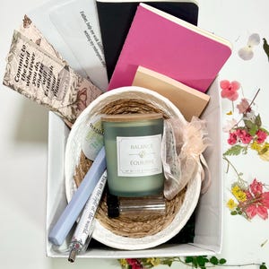Puede incluir: Un juego de regalo en una caja blanca, con una cesta tejida con una vela verde etiquetada "Balance Équilibre", cuadernos, bolígrafos y tarjetas inspiradoras con texto. El juego incluye una variedad de artículos para escribir y reflexionar.