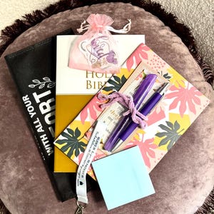May include: A Holy Bible, a black pouch with white text, a pink organza bag with stickers, a floral notebook, two purple pens tied with a purple string, and a light blue sticky note. A white lanyard with black text is draped across the items.