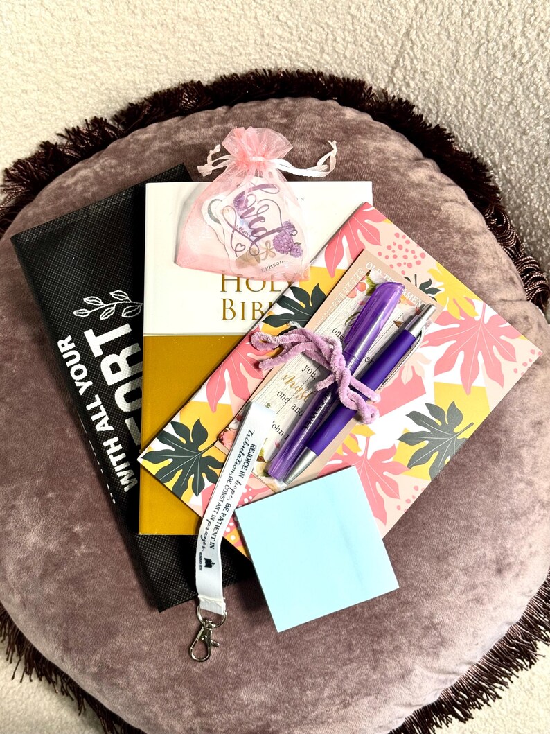 May include: A Holy Bible, a black pouch with white text, a pink organza bag with stickers, a floral notebook, two purple pens tied with a purple string, and a light blue sticky note. A white lanyard with black text is draped across the items.
