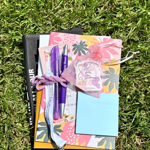 May include: A collection of stationery items including a notebook with a colourful leaf pattern, a purple pen, a purple stylus pen, a light blue sticky note pad, a white lanyard with text, and a small pink organza bag containing a sticker.