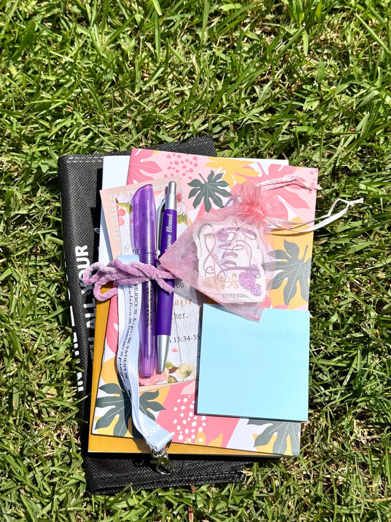 May include: A collection of stationery items including a notebook with a colourful leaf pattern, a purple pen, a purple stylus pen, a light blue sticky note pad, a white lanyard with text, and a small pink organza bag containing a sticker.