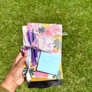 May include: A stationery set featuring a yellow notebook with a pink and green leaf pattern, two purple pens, a light blue sticky note, and a small pink organza drawstring bag containing a sticker, all tied together with a white ribbon.