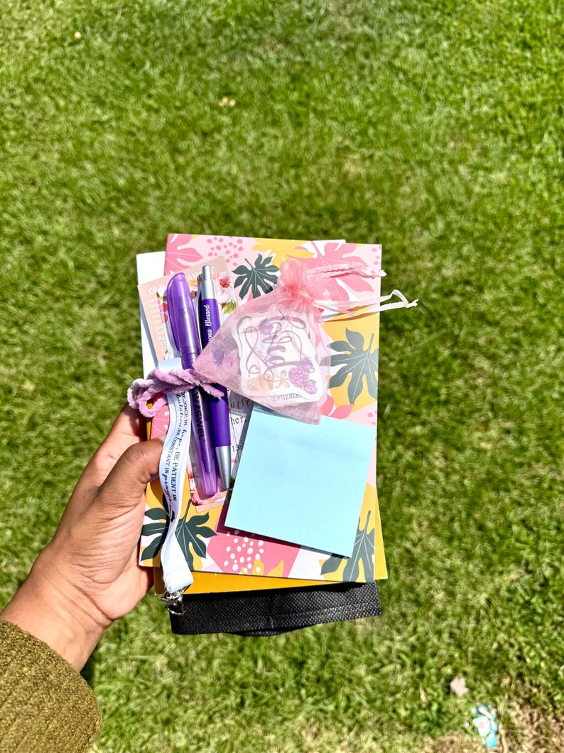 May include: A stationery set featuring a yellow notebook with a pink and green leaf pattern, two purple pens, a light blue sticky note, and a small pink organza drawstring bag containing a sticker, all tied together with a white ribbon.