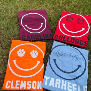 May include: Four t-shirts with a smiley face design. Each shirt has a different college mascot in the eyes of the smiley face. The shirts are red, orange, blue, and maroon. The text on the shirts reads "Carolina Bulldogs", "Clemson", "Tarheels", and "Carolina".