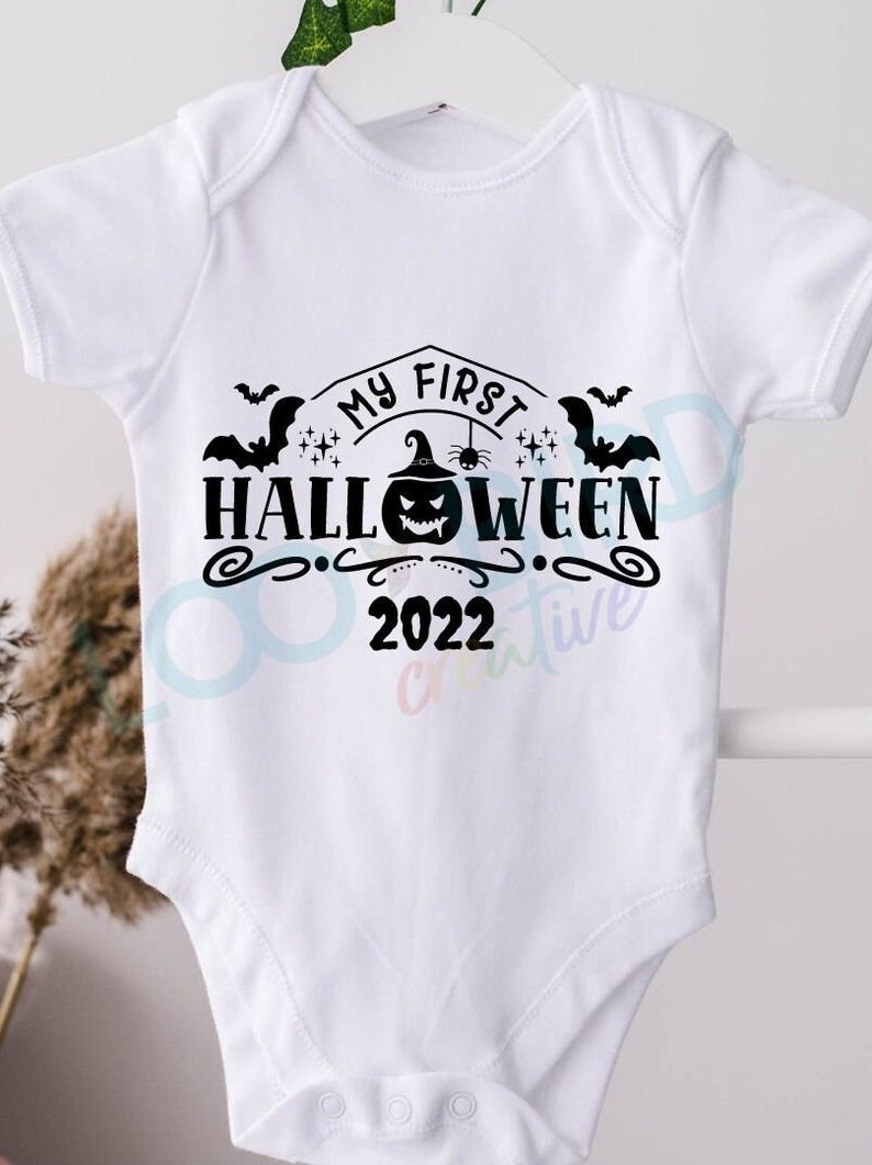 K&ouml;nnte beinhalten: Wei&szlig;er Baby-Strampler mit schwarzem Text "Mein erstes Halloween 2022" mit einem K&uuml;rbis, Flederm&auml;usen und Sternen.