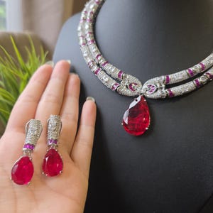 May include: A jewellery set consisting of a double-strand necklace and matching drop earrings. The necklace features silver-toned metal, clear pavé stones, and small rectangular red stones, with a large, pear-shaped red pendant at the centre.