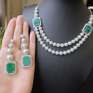 May include: A jewellery set consisting of a double-strand necklace and matching drop earrings. Each piece features rectangular green stones surrounded by small white crystals and accented with round white pearls set in a silver-toned metal frame.