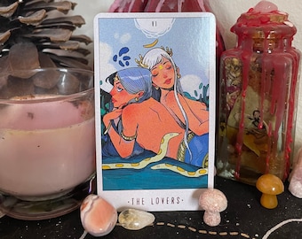 May include: A tarot card with a blue background and two figures with blue and white hair. The card is titled "The Lovers" and has the Roman numeral VI in the top right corner. The figures are holding a snake and are surrounded by water.