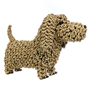 Puede incluir: Una figurita de perro tejida de color marr&oacute;n con ojos y patas de cuentas negras. El perro est&aacute; hecho de materiales naturales y tiene un aspecto r&uacute;stico.