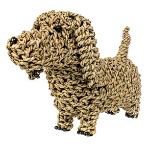 Puede incluir: Una figurita de perro de cuerda trenzada marr&oacute;n con ojos de cuentas negras. El perro est&aacute; de pie sobre sus cuatro patas con la cola enroscada.
