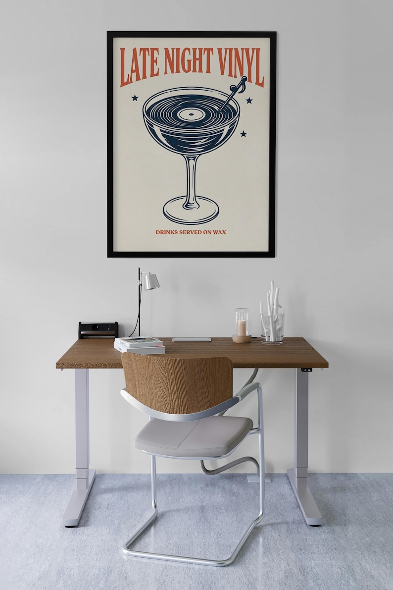 May include: A framed poster with the text "LATE NIGHT VINYL" above a cocktail glass design with a record inside. The text "DRINKS SERVED ON WAX" is below the glass. The poster is on a desk with a chair.