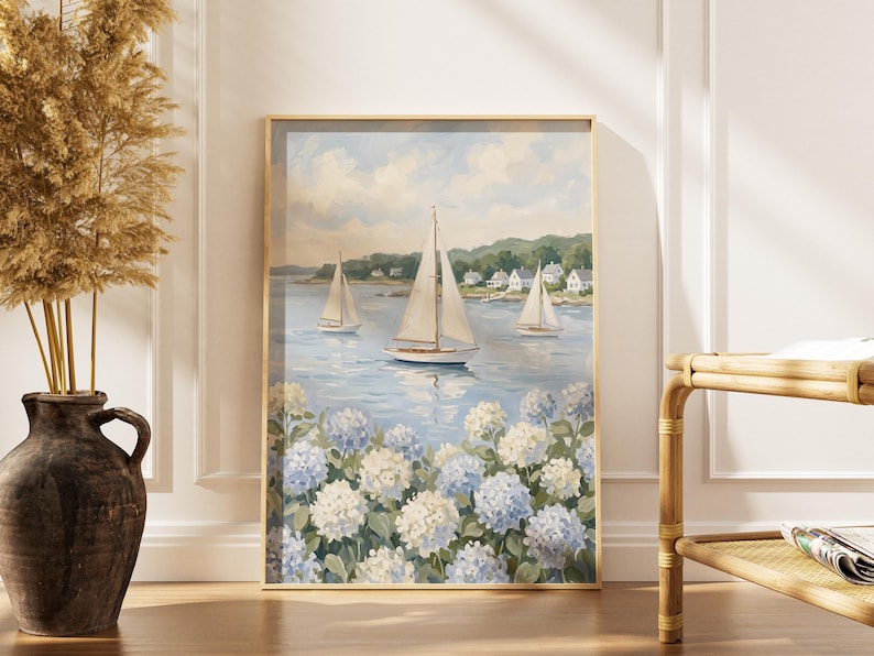 May include: A framed nautical painting depicts sailboats on a serene blue sea, with white and blue hydrangeas in the foreground. The artwork is in a gold frame, creating a coastal theme. The painting is displayed near a vase with dried plants.