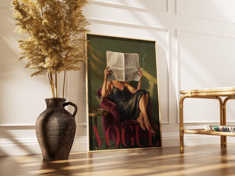 May include: A framed print featuring a woman in a black dress reading a newspaper, with the word "VOGUE" in red. The artwork has a vintage aesthetic, with a martini glass on a side table. A vase with dried plants is in the foreground.