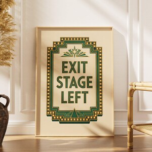 May include: A rectangular framed wall art print featuring a cream background with a dark green and gold border. The center text reads EXIT STAGE LEFT in bold green capital letters, surrounded by a decorative geometric frame with small light bulbs.
