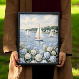 May include: A framed painting depicting sailboats on a blue sea, with white and blue hydrangeas in the foreground. The artwork features three sailboats, white houses, and a cloudy sky. The frame is black.