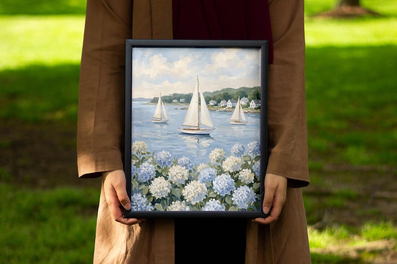May include: A framed painting depicting sailboats on a blue sea, with white and blue hydrangeas in the foreground. The artwork features three sailboats, white houses, and a cloudy sky. The frame is black.
