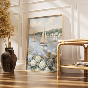 May include: A framed nautical painting featuring sailboats on a blue sea, with white and blue hydrangeas in the foreground. The painting is in a gold frame and leans against a white wall. A brown vase with dried plants and a bamboo table are in the foreground.