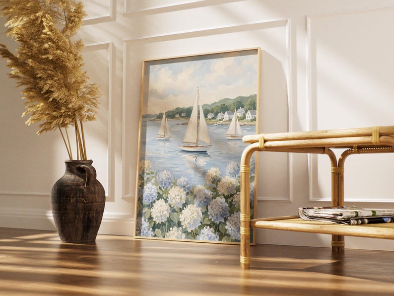 May include: A framed nautical painting featuring sailboats on a blue sea, with white and blue hydrangeas in the foreground. The painting is in a gold frame and leans against a white wall. A brown vase with dried plants and a bamboo table are in the foreground.
