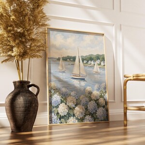 May include: A framed painting of sailboats on a blue sea, with white and blue flowers in the foreground. The painting is in a gold frame and leans against a white wall. A large brown vase with dried plants is to the left.