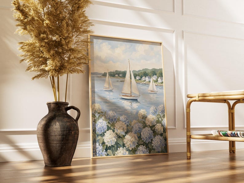 May include: A framed painting of sailboats on a blue sea, with white and blue flowers in the foreground. The painting is in a gold frame and leans against a white wall. A large brown vase with dried plants is to the left.
